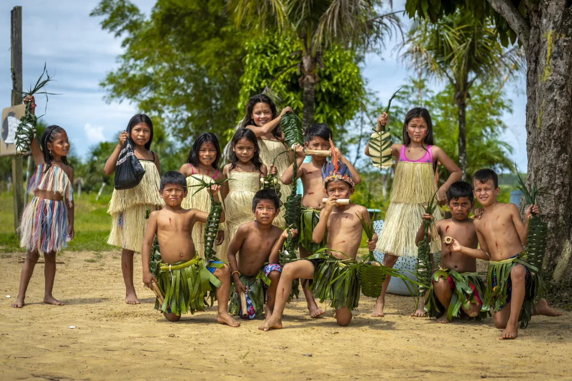 Grupo de niños y niñas del Resguardo Indígena Lagos del Paso, en Dabucury (Guaviare, Colombia), vestidos con atuendos tradicionales hechos de fibras y hojas, posando al aire libre y sosteniendo elementos elaborados con palma.