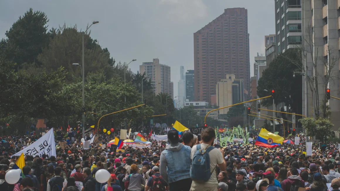 Crowd moving through a Bogotá avenue during a nighttime demonstration, surrounded by red traffic lights, urban buildings and dense city atmosphere, reflecting political tension and social uncertainty in Colombia ahead of the elections.