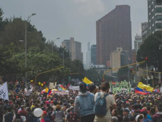 Crowd moving through a Bogotá avenue during a nighttime demonstration, surrounded by red traffic lights, urban buildings and dense city atmosphere, reflecting political tension and social uncertainty in Colombia ahead of the elections.